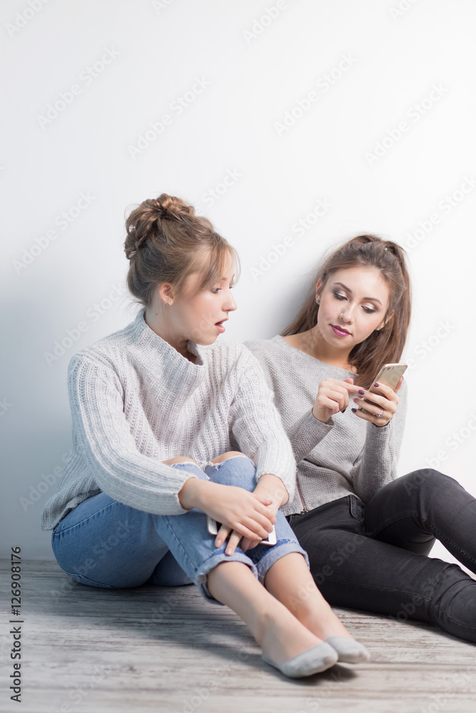 portrait two surprised girls looking at cell phone discussing latest gossip news, shopping, shocked at what they see, isolated white background