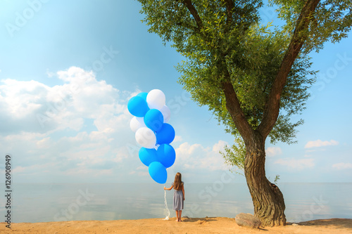 Little girl stands on the shore near the tree with balloons in h
