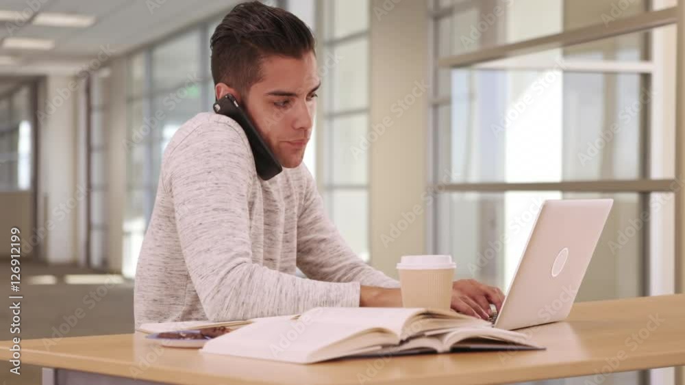 Millennial Latino university student in campus library with laptop and cellphone. Young Hispanic man studying and doing homework in public space Millennial Latino university student in campus library with laptop and cellphone. Young Hispanic man studying and doing homework in public space