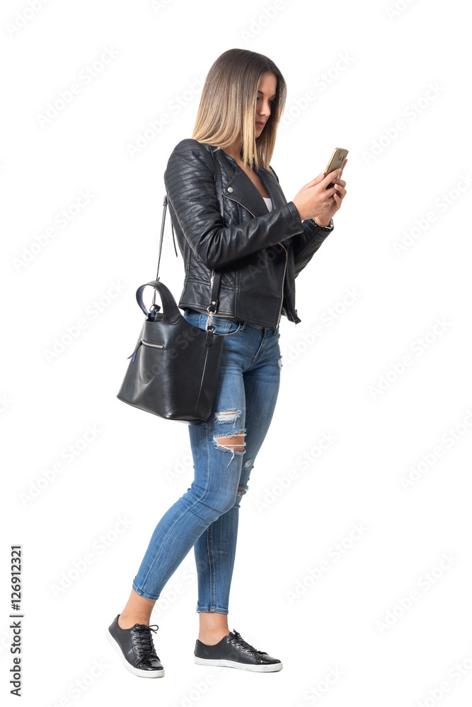 Fototapeta premium Casual young woman in street style clothing walking and typing on mobile phone. Full body length portrait isolated over white background