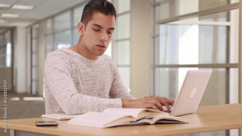 Millennial Hispanic university student in campus library with laptop and cellphone. Young Latino man studying and doing homework in public space Millennial Hispanic university student in campus library with laptop and cellphone. Young Latino man studying and doing homework in public space