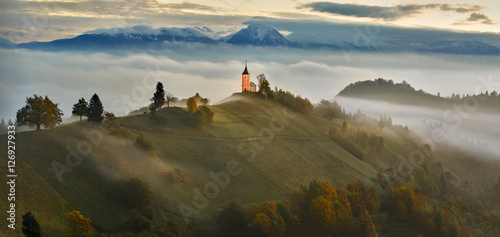 In the morning mist, St. Primoz church near Jamnik at dusk with alps in background, Slovenia