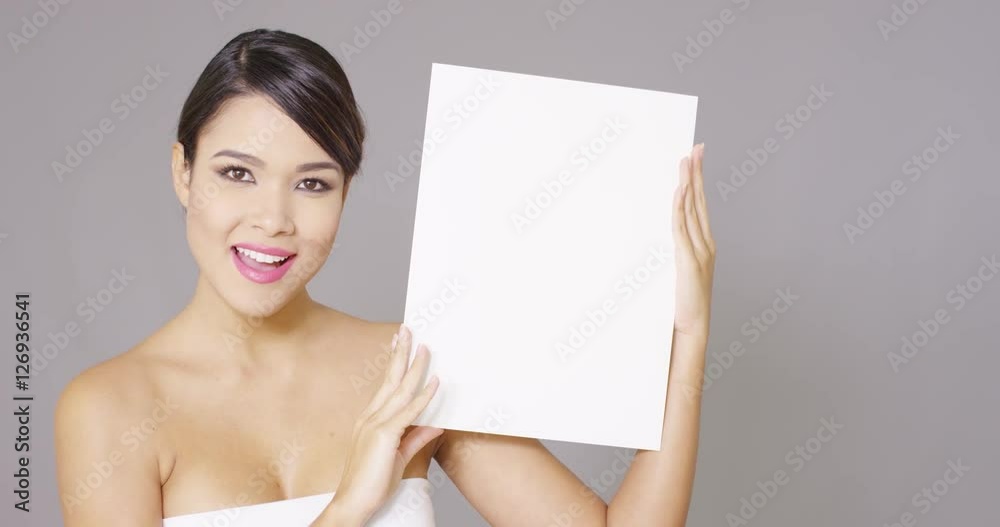 Young woman displaying a blank white sign to the side of her body with a charming friendly smile over grey Young woman displaying a blank white sign to the side of her body with a charming friendly smile over grey