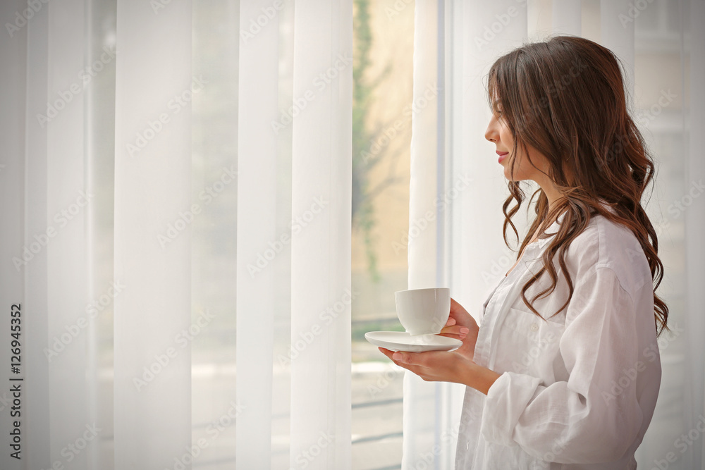Beautiful woman with cup of aromatic coffee standing near window Beautiful woman with cup of aromatic coffee standing near window