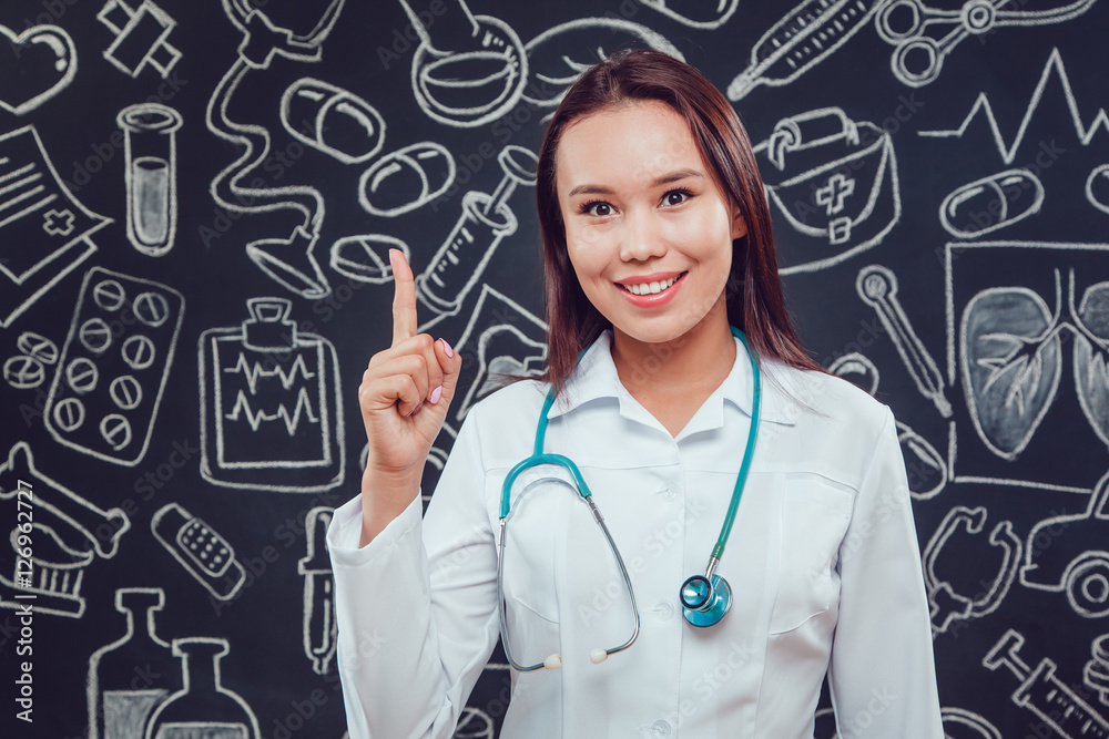 Smiling young woman doctor in white coat standing on dark background with pattern. She raised her thumb up