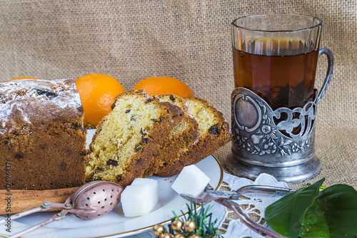 Cake with raisins and black tea in glass with glass holder, vintage tea strainer, pinces for sugar on sackcloth background photo