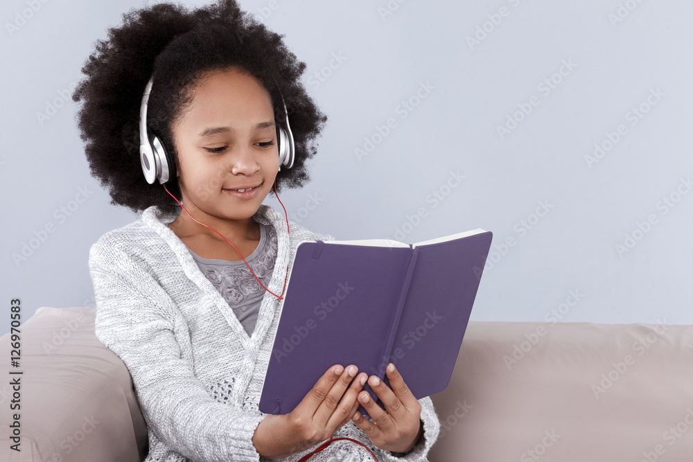 Pretty little girl in headphones holding notebook and smiling while sitting on sofa Pretty little girl in headphones holding notebook and smiling while sitting on sofa