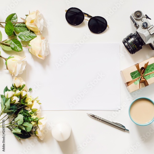 White desk with coffee cup  sunglasses  roses and film camera. Empty sheet in the middle. Top view  flat lay  copyspace.