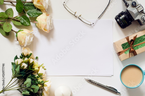 White desk with coffee cup, sunglasses, roses and film camera. Empty sheet in the middle. Top view, flat lay, copyspace.