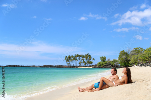 Beach vacation relaxation. Happy couple relaxing sunbathing lying down on sand together tanning under the tropical sun in getaway travel destination. Young adults enjoying their holidays.