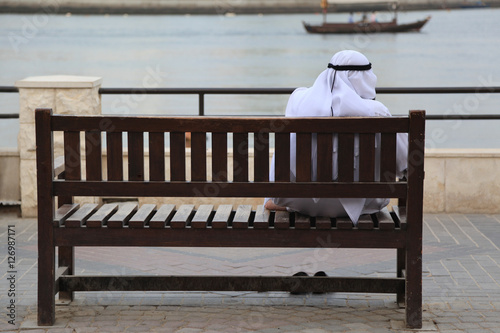 Arab man in dishdasha sitting on a wooden bench at Creek, Dubai photo