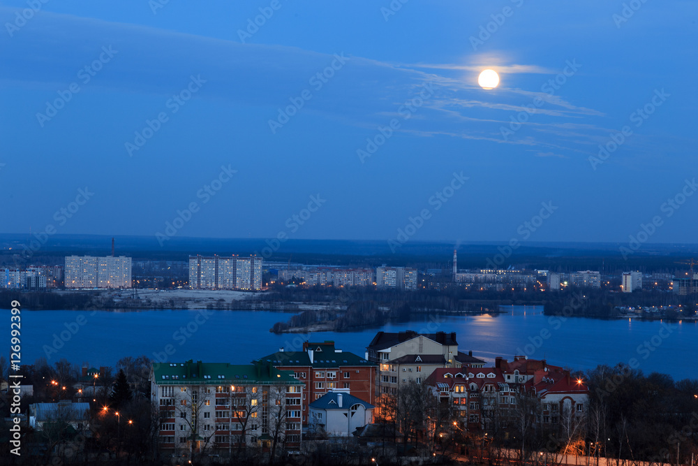 View of early moon in blue evening sky with reflection in the river. Cityscape 