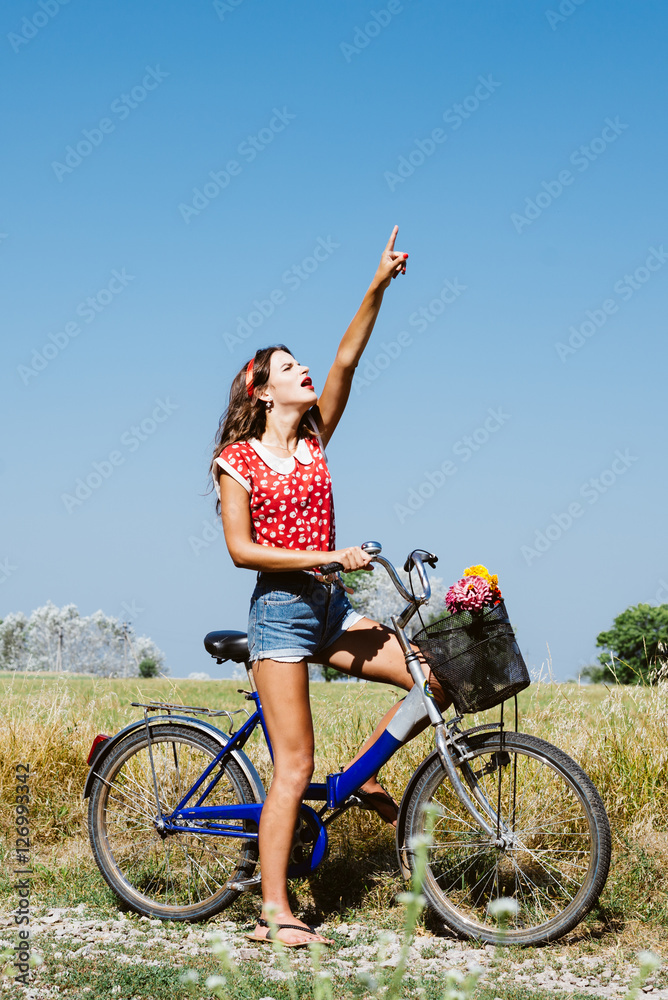 Beautiful pinup girl having fun cycling in fields under bright blue summer sky and pointing up on outdoors copy space background Beautiful pinup girl having fun cycling in fields under bright blue summer sky and pointing up on outdoors copy space background