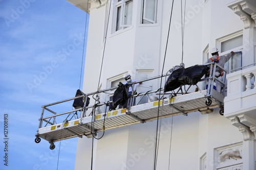 High above the ground, scaffolding workers repaint an apartment building. Blue sky. Horizontal.