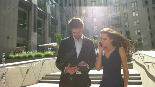 Two young colleagues walking on stairs, woman talking on phone, man using tablet outdoors.