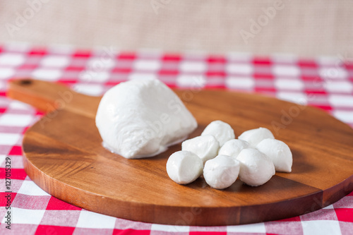 Water buffalo mozzarella mozzarella di buffala cheese in ball and bocconcini balls on wooden cheese chopping board on red and white checkered tableclothbrown burlap background copy space above