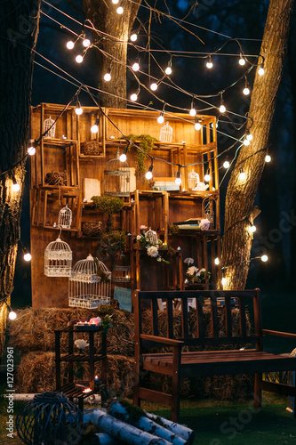 Beautiful illumination on rustic designed wall. Rack with wicker baskets, cages and haystack, lighted with garland. Evening, romantic decoration, creative design concept