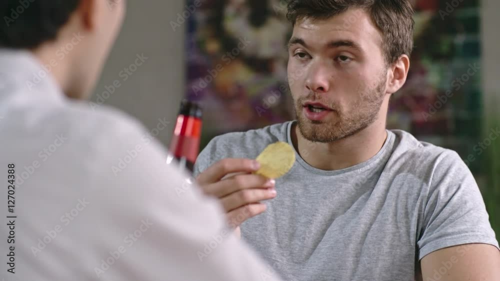Over the shoulder shot of tipsy man with stubble eating potato chips and talking with friend at party Over the shoulder shot of tipsy man with stubble eating potato chips and talking with friend at party