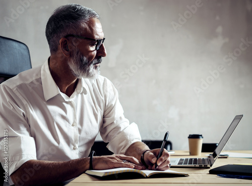 Adult successful businessman wearing a classic glasses and working with laptop at the wood table in modern loft.Bearded middle age man making notes notebook. Horizontal,blurred background.