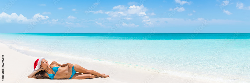 Christmas vacation beach woman banner panorama. Wide paradise beach background with Asian bikini woman lying down relaxing on white sand beach with christmas santa hat.