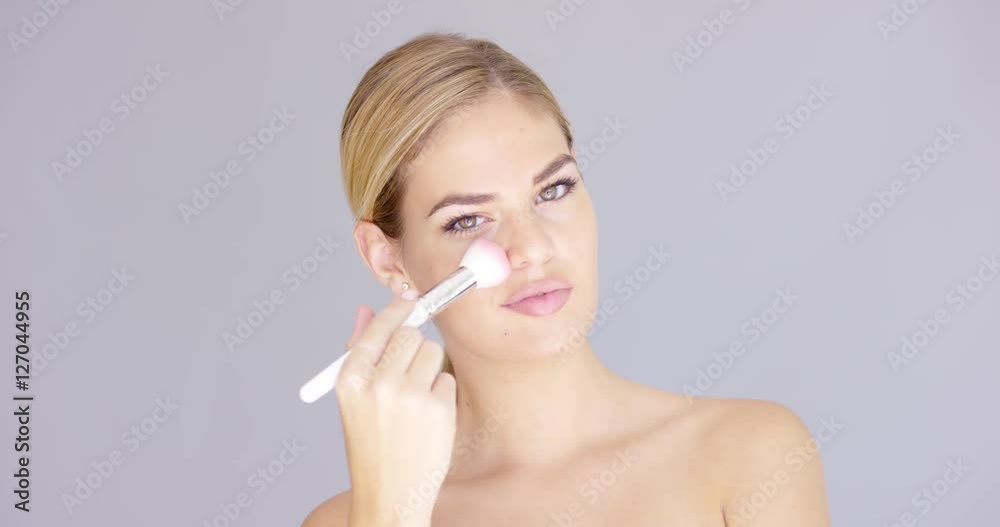 Beautiful young female applying makeup on her cheek with cosmetic brush. Portrait with bare shoulders over isolated gray background. Looking at camera Beautiful young female applying makeup on her cheek with cosmetic brush. Portrait with bare shoulders over isolated gray background. Looking at camera