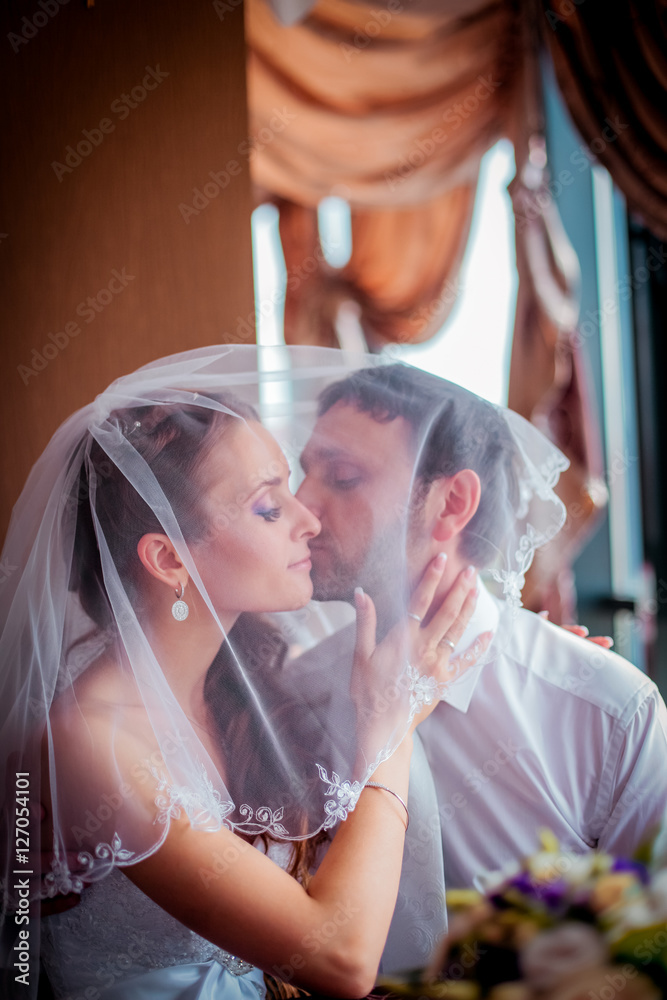 Fototapeta premium Bride kisses on the cheek the groom happy couple holding a bouquet in their hands in the dark interior near the window. Close-up
