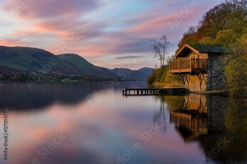 Pink and purple vibrant sunrise with dawn light hitting boathouse on calm lake with reflections in the Lake District. © _Danoz