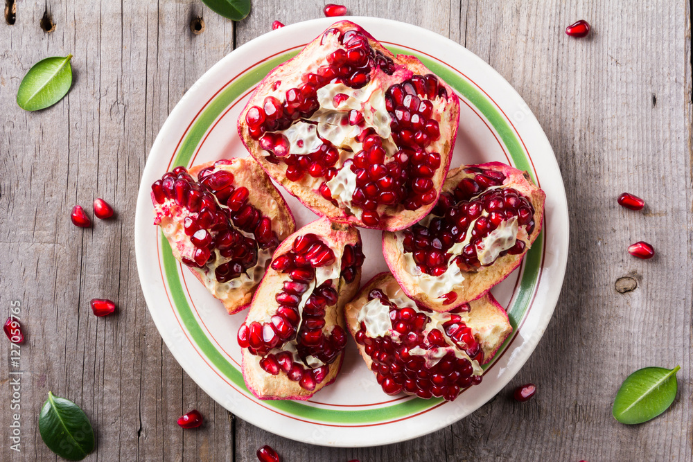 Broken red ripe juicy pomegranates in the white plate on rustic wooden unpainted table Broken red ripe juicy pomegranates in the white plate on rustic wooden unpainted table