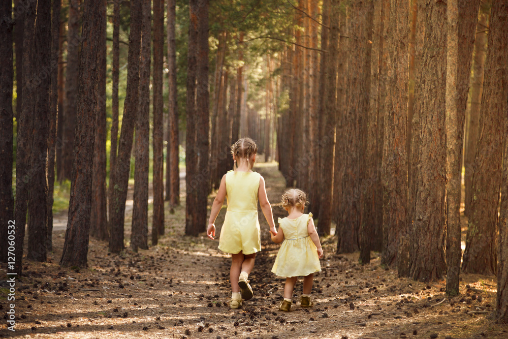 Fototapeta premium Two sisters in yellow dresses go through the forest holding hand