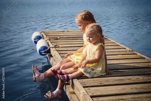 Two little sisters in yellow dresses sitting at wooden pier near