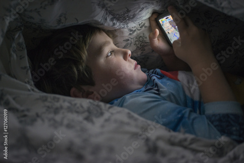 Young boy playing with cell phone under duvet