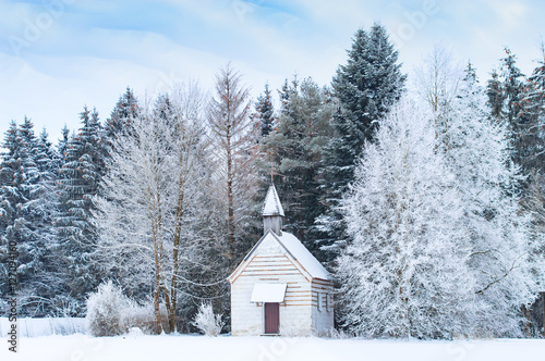 Small wooden chapel on snowbound frosty glade in snowy frozen forest. Winter wonderful scenery in German Bavarian region Allgaeu at the foot of Alps.