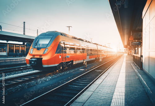 Modern high speed red commuter train at the railway station at colorful sunset. Railroad with vintage toning. Train at railway platform. Industrial landscape. Railway tourism. Vintage toning. Concept