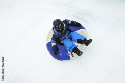 A child boy (wearing ski helmet) having fun sledding on a tube in the snow. 