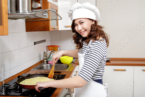 Beautiful Asian woman in white chef hat preparing an omelet in the kitchen. Looking at camera.