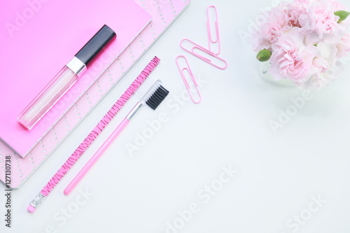 A feminine pink desktop mock up with pink lipstick, notebooks, paper clips, a pencil and miniature pink carnations on a white isolated background for a business concept.