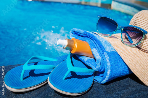 Blue slippers with sunscreen cream, towel, straw hat and sunglasses on border of a swimming pool - holiday tropical concept