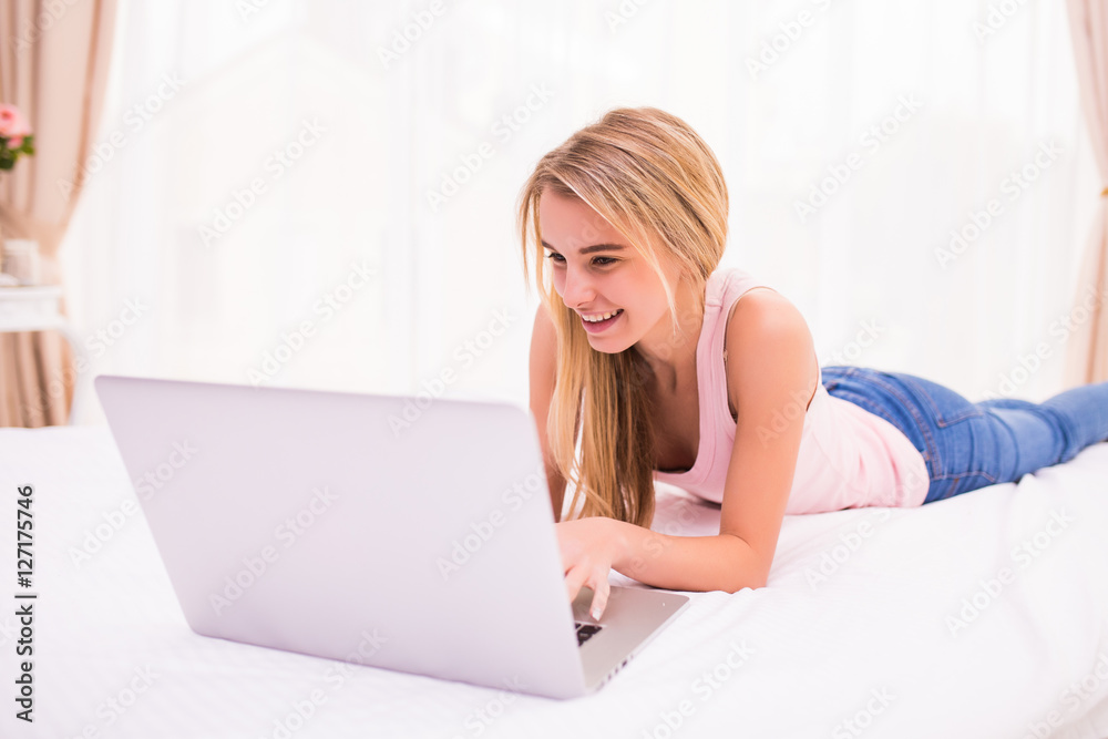 Cheerful attractive young woman lying on bed and using laptop