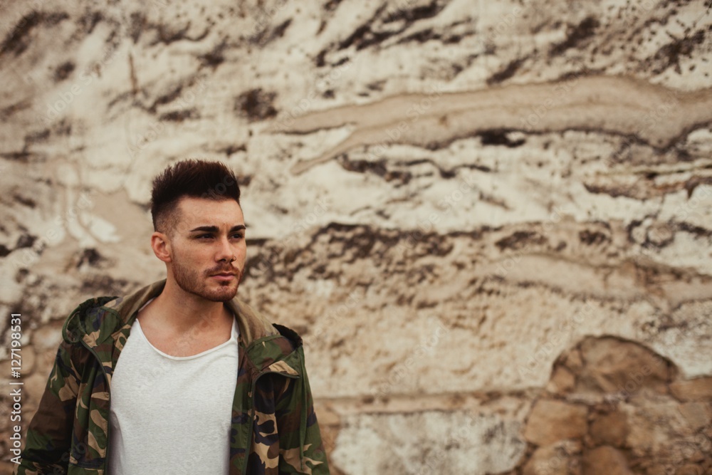 Attractive guy with jacket military stylish on a wall of mud