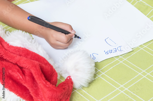 Children's hand is writing a letter to Santa Claus. On the table is Santa's cap.