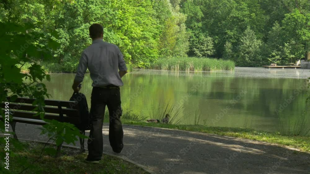 Man Comes to Rest in Park Looking at Wild Ducks Sits Down to the Bench by Lake Fresh Green Trees Sunny Spring Day Smooth Water Tourist on Wooden Bench Man Comes to Rest in Park Looking at Wild Ducks Sits Down to the Bench by Lake Fresh Green Trees Sunny Spring Day Smooth Water Tourist on Wooden Bench