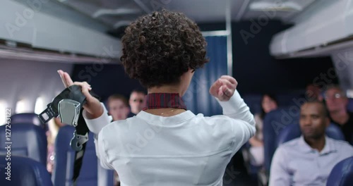 Commercial airline passengers watch as female flight attendant points out emergency exits during safety briefing.  Medium shot from front of plane recorded at 60fps