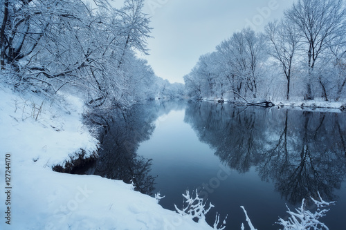 Winter forest on the river at sunset. Colorful landscape with snowy trees, beautiful frozen river with reflection in water. Seasonal. Winter trees, lake and blue sky. Frosty snowy river. Weather
