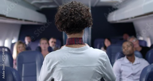 Commercial airline passengers watch as female flight attendant demonstrates seat belt during safety briefing.  Medium shot from front of plane with camera dolly.
