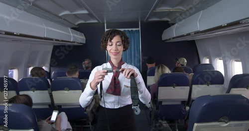 Female flight attendant demonstrates seat belt to passengers in main cabin of commercial airliner.  Medium long shot with camera dolly.