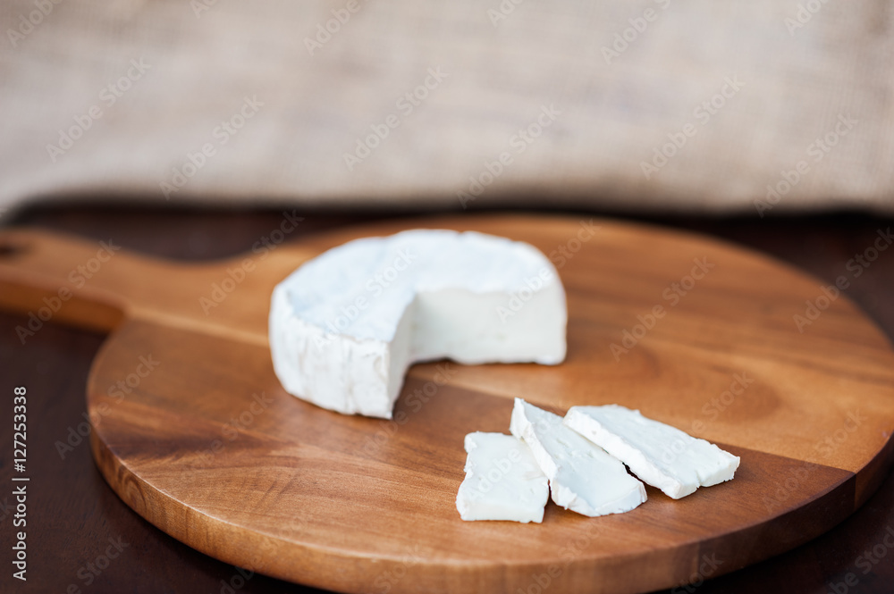 Brie or camembert cheese cut out wedge and in slices on a wooden chopping cutting cheese board on dark brown wooden table with a light brown burlap background
