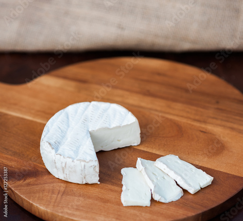Brie or camembert cheese cut out wedge and in slices on a wooden chopping cutting cheese board on dark brown wooden table with a light brown burlap background