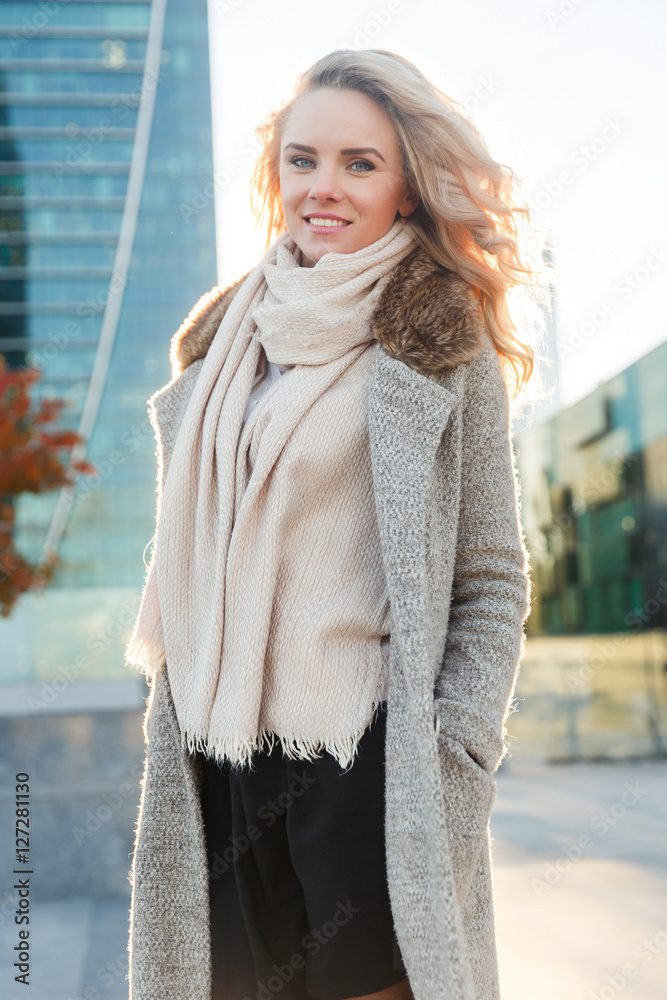 Young blond woman in long coat standing among buildings Young blond woman in long coat standing among buildings