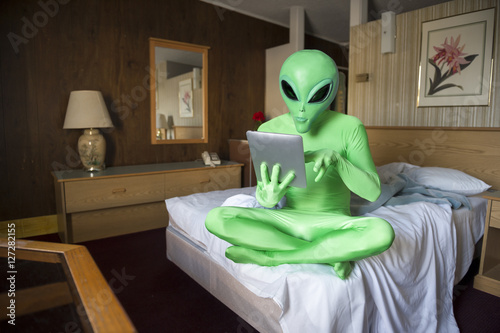 Green alien sitting using futuristic tablet computer on the bed in an old-fashioned room