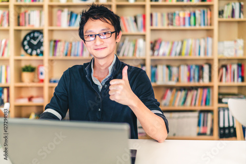 Obraz na plátně Young adult Asian man with laptop, thumbs up ok sign, home office or library sce